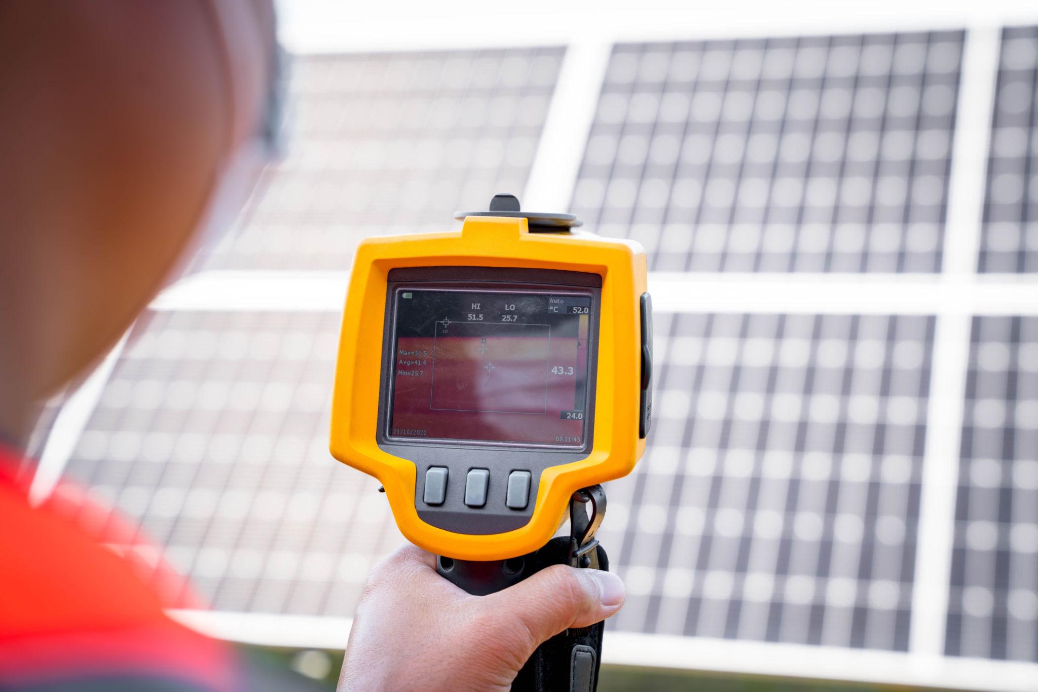Person holds an orange infrared thermal camera up to solar panels, inspecting for hotspots among the panels