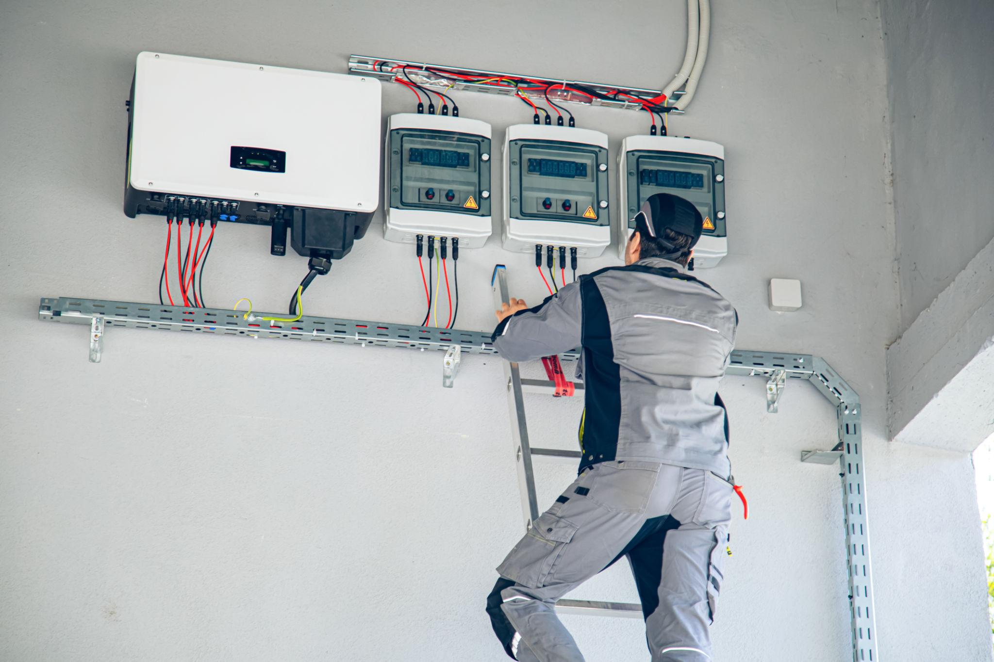 Electrician on a ladder inspecting electrical meters mounted on a wall in an industrial setting