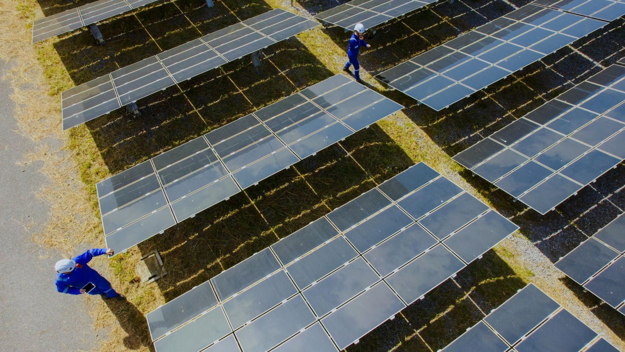 Bird's-eye view of a solar farm with rows of photovoltaic panels and two workers in blue coveralls and white helmets inspecting them.