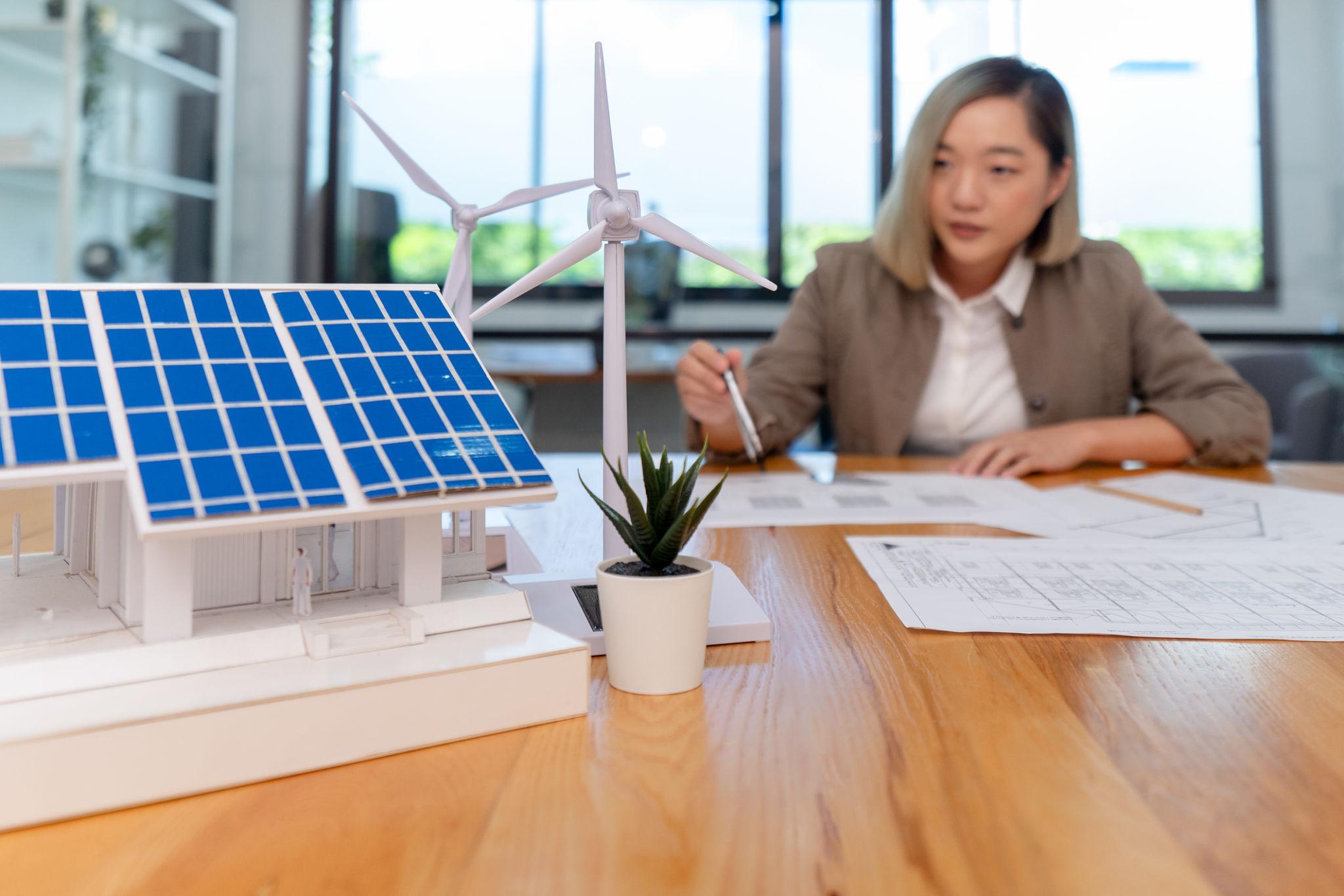 Businesswoman reviewing blueprints at a table with solar panel and wind turbine models nearby on a wooden surface.