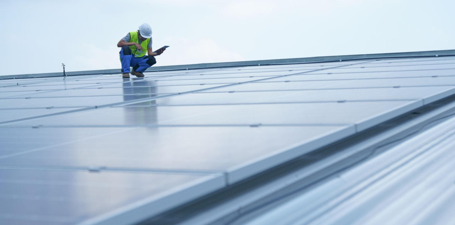 Construction worker in a safety vest and helmet kneels on a solar panel roof, inspecting panels with a tablet.